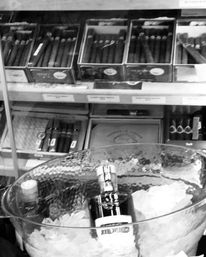 Chilled liquor bottle in a glass ice bucket on a counter with rows of boxed cigars on shelves behind — cigar shop display, tobacco store retail scene.