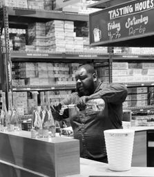 Person pouring a spirits sample at a liquor tasting counter in a warehouse-style retail store with boxed shelves and a chalkboard listing tasting hours.