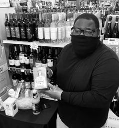 Masked store associate showcasing a boxed tequila bottle at a liquor-store tasting table with wine bottles lining the shelves.