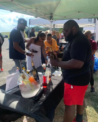 Outdoor summer tasting under pop-up tents with people sampling wine and spirits — server opening a bottle beside a table of chilled bottles in an ice bucket and a small crowd.