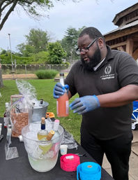 Gloved bartender pouring a pink cocktail at an outdoor park event, ice bucket with wine bottles, rolls of raffle tickets on the table and a wooden pavilion in the background.