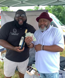 Two smiling men under a vendor canopy at an outdoor market — one holds a wine bottle, the other fans cash, with boxes of bottles and a white van in the background.