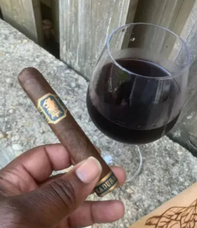 Close-up of a hand holding a Maduro cigar beside a glass of red wine on a concrete patio ledge with a weathered wooden fence background.