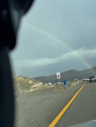 Rainbow arching over a highway through dry hills under a stormy sky, cars in traffic and a no U-turn sign beside a yellow-lined road.