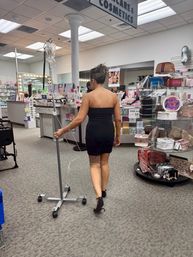 Woman in a black strapless dress and heels walking through a cosmetics store while rolling an IV pole past makeup counters and accessory displays.
