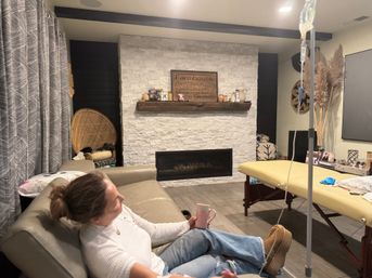 Cozy farmhouse-style living room with white stone fireplace and rustic mantel sign, person relaxing on a leather sofa with a pink mug, rattan chair and patterned curtains, portable massage table and IV drip stand in the foreground, pampas grass decor.