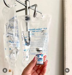 Hand with polished nails holding a small vial labeled CBD in front of clear IV saline bags hanging from a chrome IV pole in a clinical setting.