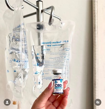 Hand with polished nails holding a small vial labeled CBD in front of clear IV saline bags hanging from a chrome IV pole in a clinical setting.
