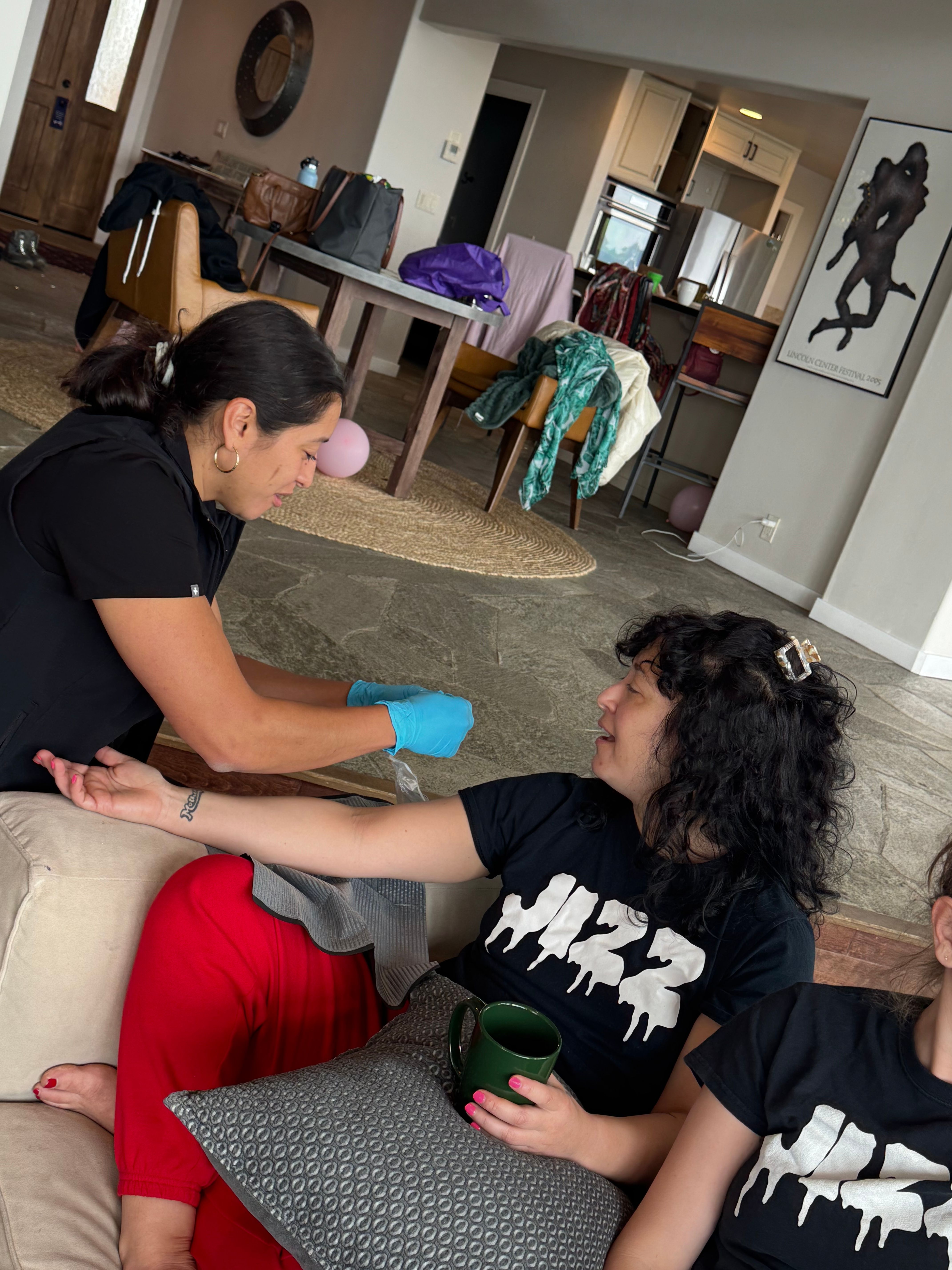 Cozy home scene: caregiver wearing blue gloves draws blood from a relaxed woman on a sofa holding a green mug, with dining table and jackets in the background.