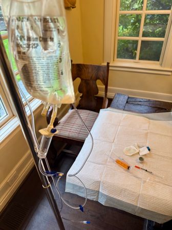 At-home infusion setup: IV bag and clear tubing on a metal pole beside a windowed dining table, with a disposable underpad, syringe, alcohol swabs and bandages on the table.