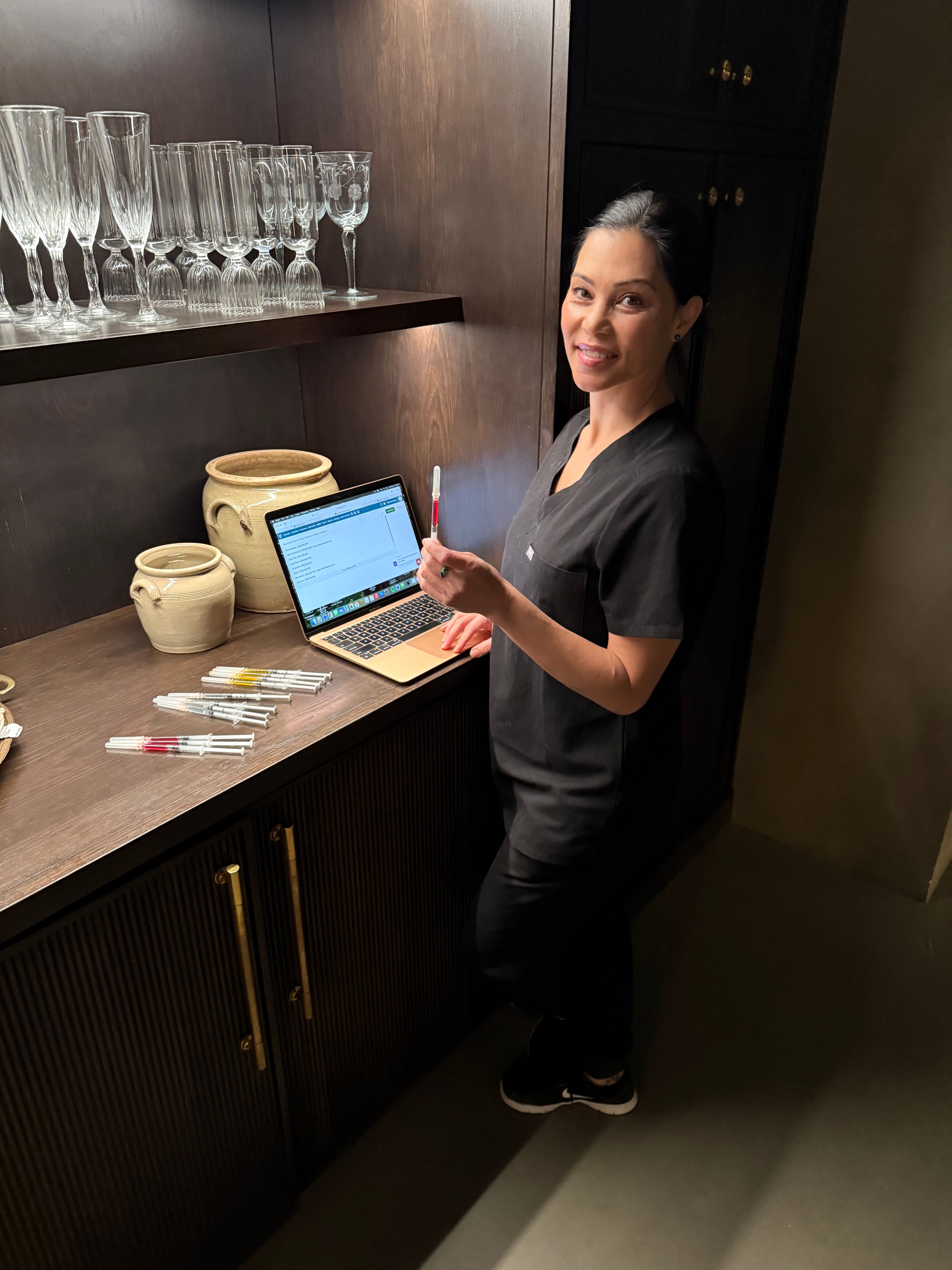 Smiling clinician in black scrubs at a dark wood treatment counter with an open laptop, holding a filled syringe while several syringes, ceramic jars and glassware sit on the shelf in a dimly lit treatment room