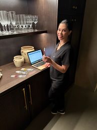 Smiling clinician in black scrubs at a dark wood treatment counter with an open laptop, holding a filled syringe while several syringes, ceramic jars and glassware sit on the shelf in a dimly lit treatment room