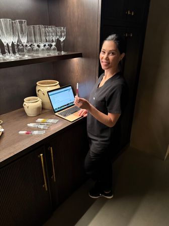 Smiling clinician in black scrubs at a dark wood treatment counter with an open laptop, holding a filled syringe while several syringes, ceramic jars and glassware sit on the shelf in a dimly lit treatment room