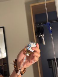 Hand with wooden bead bracelet drawing liquid from a CBD vial into a syringe beside an IV pole and open closet in a room