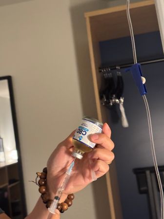 Hand with wooden bead bracelet drawing liquid from a CBD vial into a syringe beside an IV pole and open closet in a room