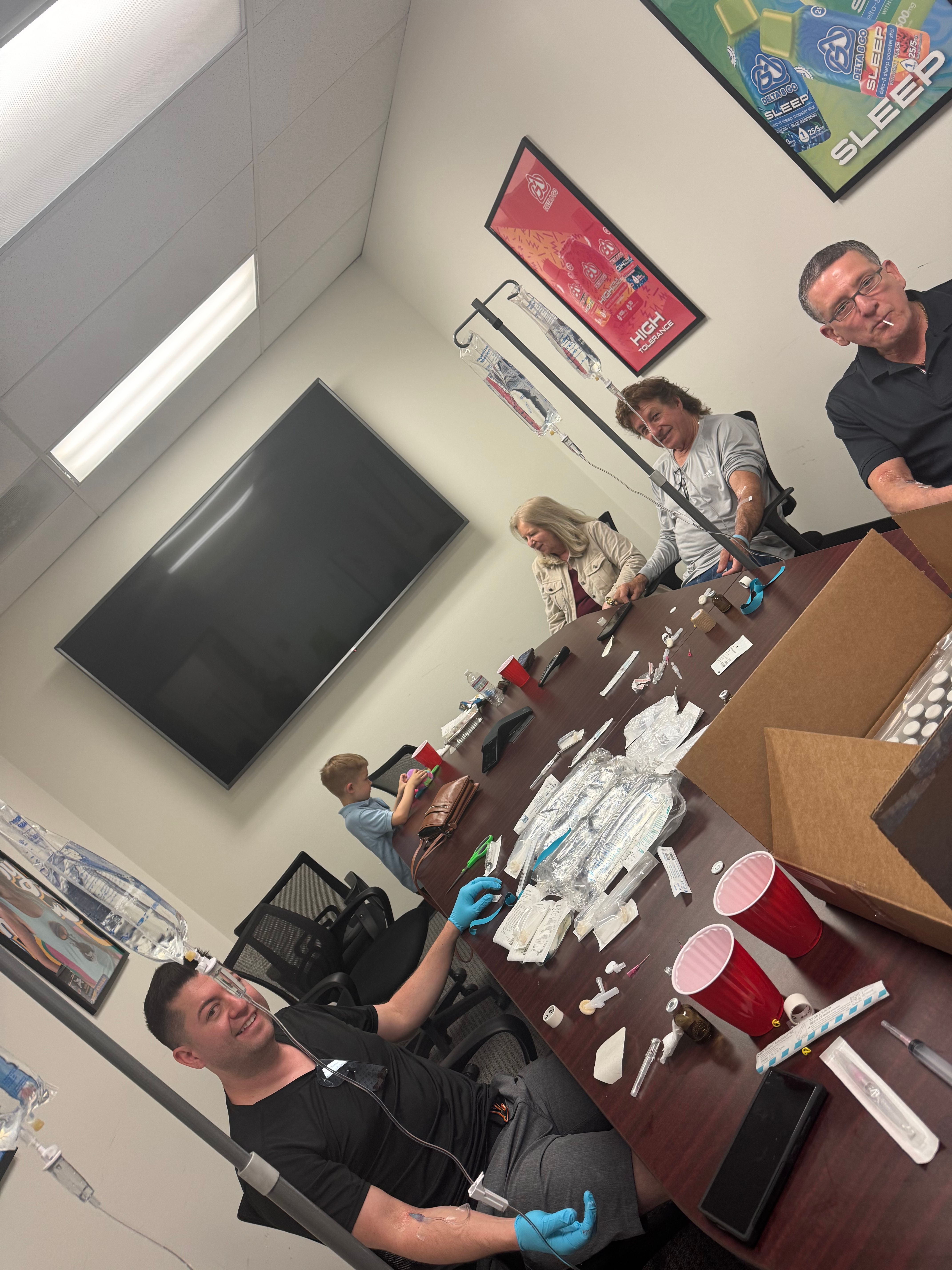 Group IV therapy session in a clinic-style conference room — adults and a child seated around a table with IV poles, infusion bags, medical supplies and red cups.