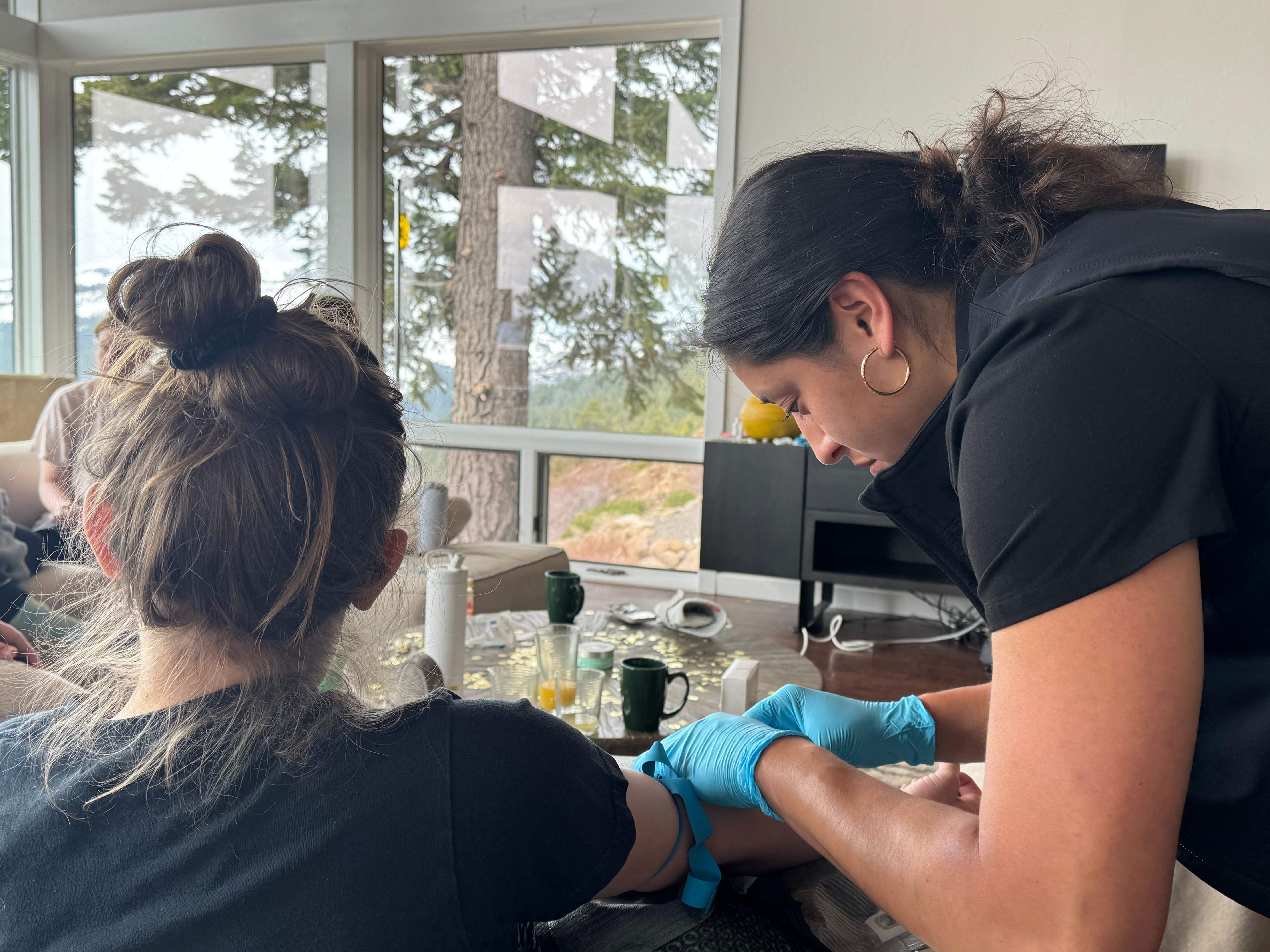Healthcare worker wearing blue gloves prepares an at-home blood draw, fastening a tourniquet on a seated person's arm in a bright living room with large windows overlooking pine trees, with mugs and medical supplies on the coffee table.
