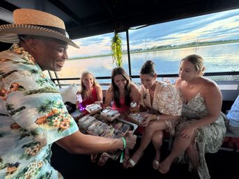Smiling man in a straw hat and tropical shirt offers trays of boxed sushi to four women seated together on a sunset boat cruise over calm water and distant shoreline.