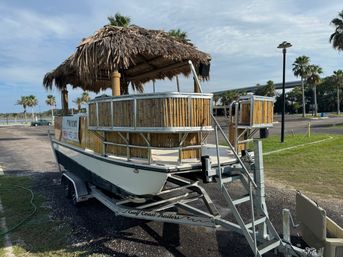 Tiki-style pontoon boat with thatched roof and bamboo railings parked on a trailer in a waterfront lot, palm trees and a bridge in the background, 'For Rent' sign visible.