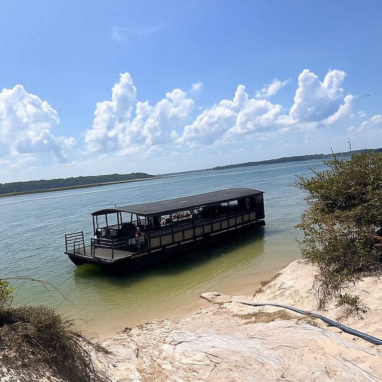 Covered sightseeing boat moored along a sandy rocky shoreline with shrubs on a calm coastal lagoon beneath blue sky and puffy white clouds.