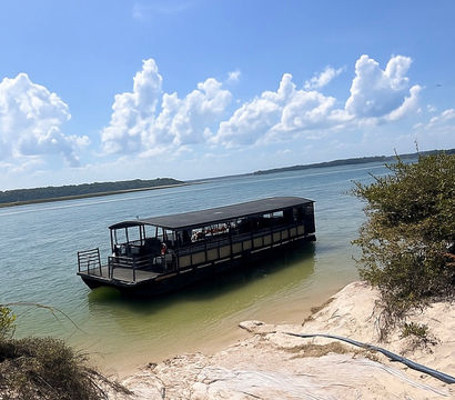 Covered sightseeing boat moored along a sandy rocky shoreline with shrubs on a calm coastal lagoon beneath blue sky and puffy white clouds.