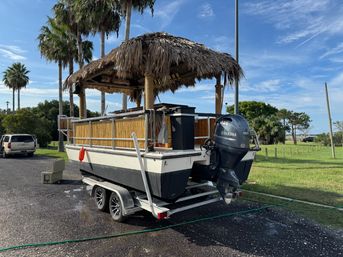 Tiki-style pontoon boat with thatched roof and bamboo railings on a double-axle trailer, parked under palm trees in a coastal park with a visible outboard motor.