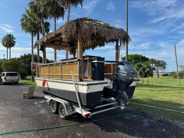 Tiki-style pontoon boat with thatched roof and bamboo railings on a double-axle trailer, parked under palm trees in a coastal park with a visible outboard motor.