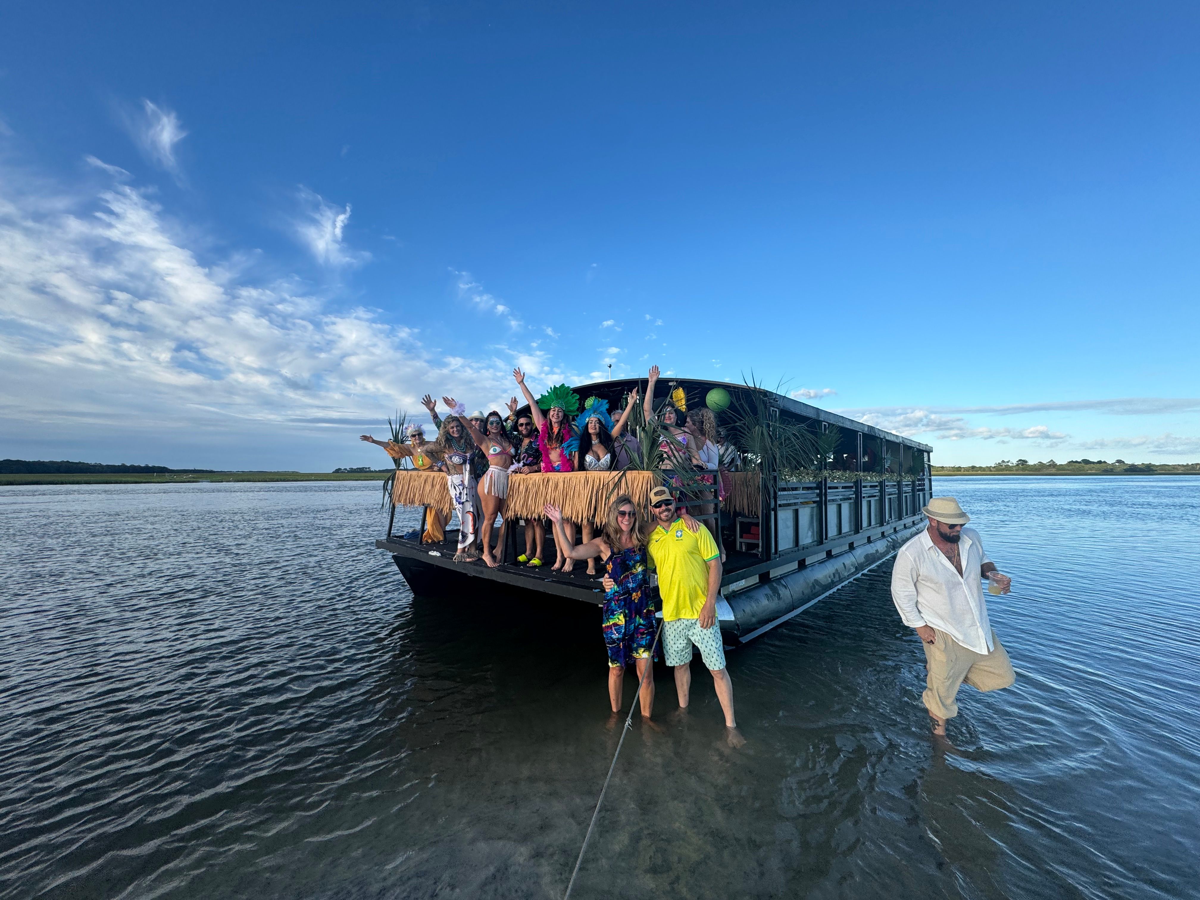 Tiki-themed pontoon boat party anchored in shallow coastal inlet, group waving from a grass-skirted deck with palm decor under a bright blue sky.