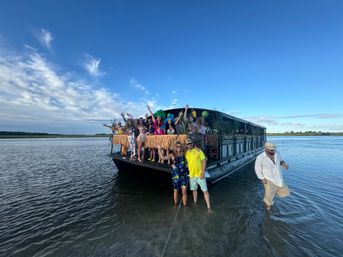 Tiki-themed pontoon boat party anchored in shallow coastal inlet, group waving from a grass-skirted deck with palm decor under a bright blue sky.