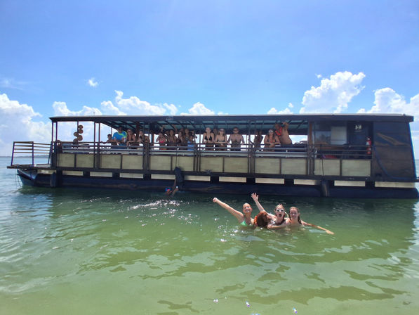 People swimming and cheering in green-blue coastal water beside a covered party boat under a bright blue sky with puffy clouds