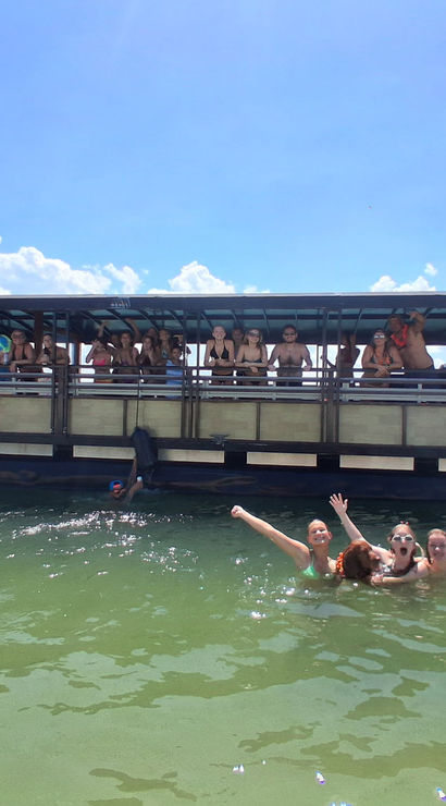 People swimming and cheering in green-blue coastal water beside a covered party boat under a bright blue sky with puffy clouds