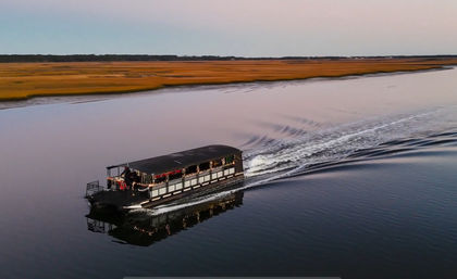 Covered pontoon boat trimmed with string lights cruising a calm tidal creek at sunset, leaving rippled reflections by golden coastal marsh grasses.