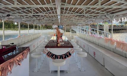 Interior of a white party pontoon boat with a thatched canopy, central high table surrounded by round stools, long bench seating along both sides, pink tassel decorations and a 'HAPPY' banner, docked by a sandy waterfront