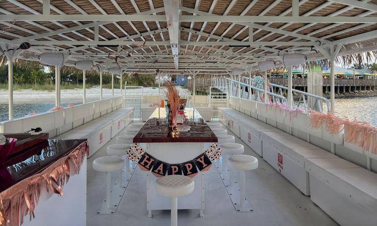 Interior of a white party pontoon boat with a thatched canopy, central high table surrounded by round stools, long bench seating along both sides, pink tassel decorations and a 'HAPPY' banner, docked by a sandy waterfront
