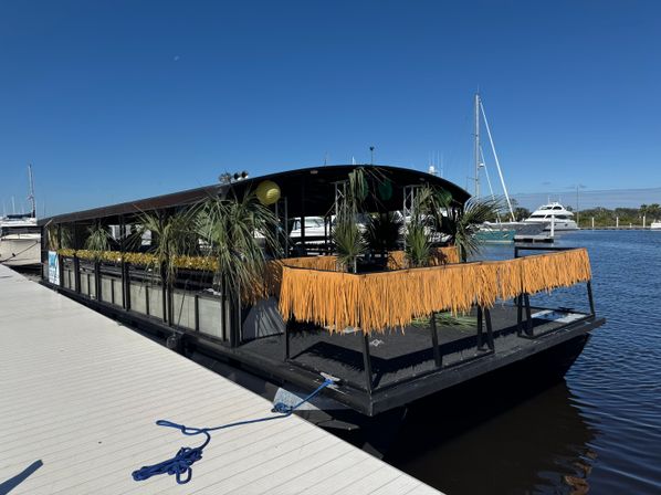 Tiki-themed floating bar docked at a sunny marina with palm plants, orange fringe trim, nearby yachts and clear blue sky.