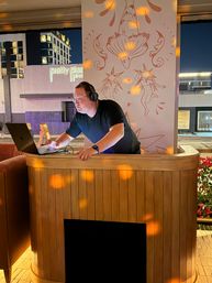 Nighttime rooftop lounge DJ with headphones and glasses leaning over a laptop behind a curved wooden booth, warm disco light spots and star-themed mural on the wall, city buildings visible outside.