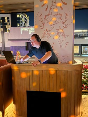 Nighttime rooftop lounge DJ with headphones and glasses leaning over a laptop behind a curved wooden booth, warm disco light spots and star-themed mural on the wall, city buildings visible outside.