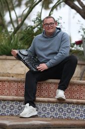Person in a gray hoodie and glasses sitting on mosaic-tiled outdoor steps holding a portable DJ controller, white sneakers visible — casual, ready to spin.