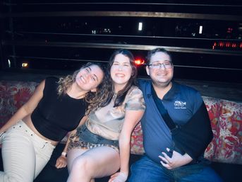 Three friends smiling together on a patterned bench at a rooftop bar at night with city lights in the background.