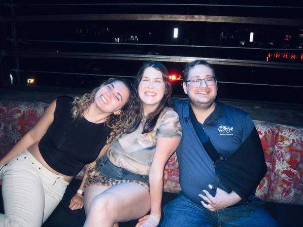 Three friends smiling together on a patterned bench at a rooftop bar at night with city lights in the background.