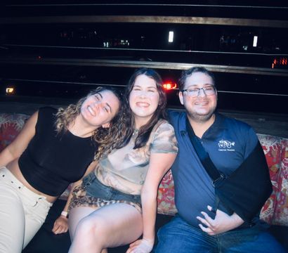 Three friends smiling together on a patterned bench at a rooftop bar at night with city lights in the background.