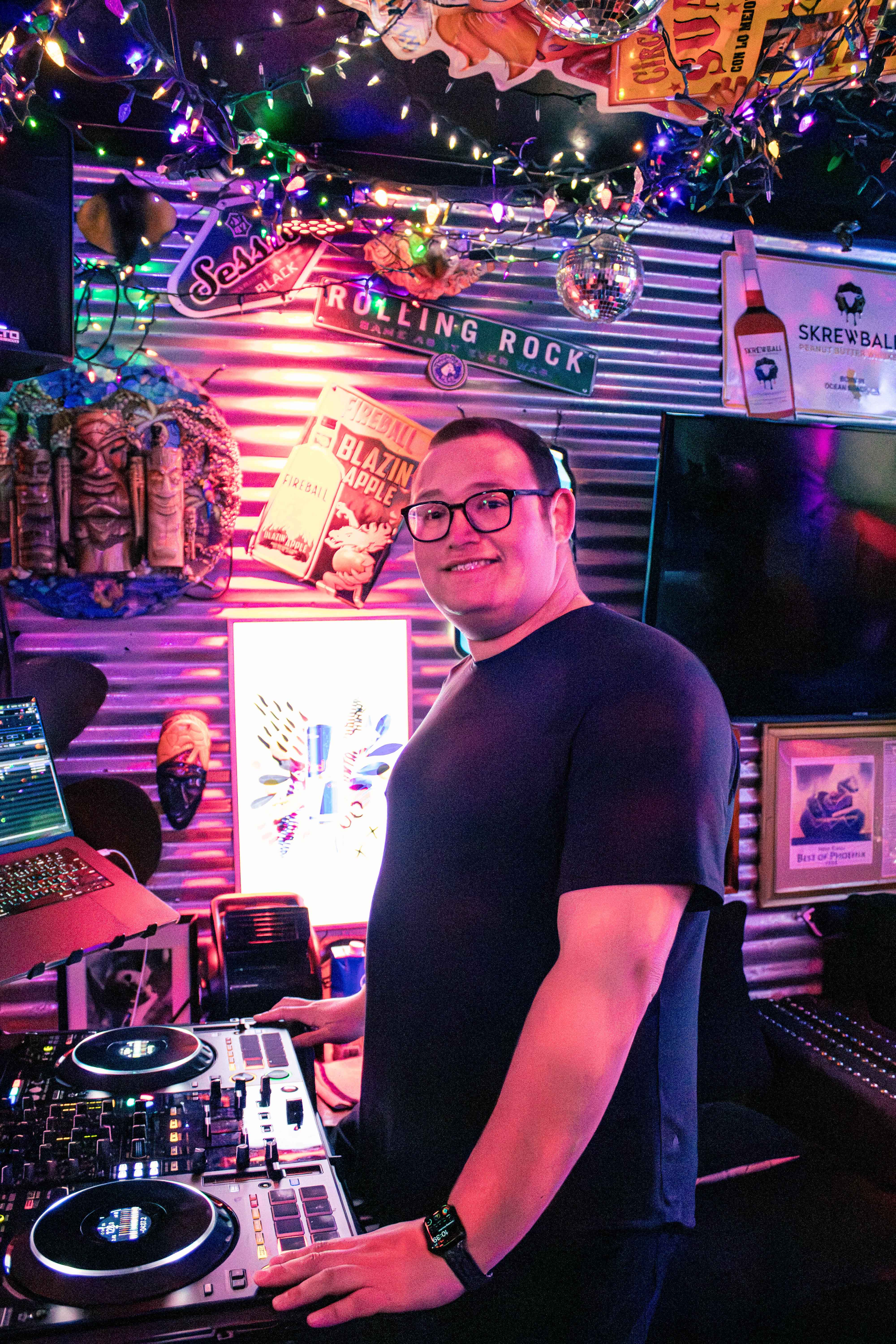 DJ at a neon-lit bar smiling behind a DJ controller and laptop, surrounded by colorful string lights, disco ball and eclectic wall decor
