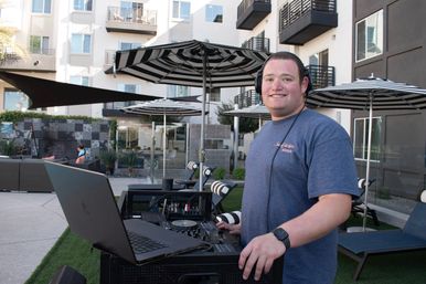 DJ with headphones working a laptop and mixer in a modern apartment courtyard outdoor lounge with black-and-white striped umbrellas and lounge chairs