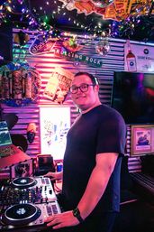 Smiling DJ with glasses mixing on turntables in a neon-lit bar, surrounded by string lights, disco balls, tiki masks and vintage drink signs