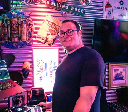 Smiling DJ with glasses mixing on turntables in a neon-lit bar, surrounded by string lights, disco balls, tiki masks and vintage drink signs
