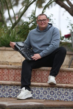 Smiling person in a gray hoodie and glasses sitting on patterned outdoor tiled steps, holding a DJ controller and wearing black jeans and white sneakers, casual DJ vibe.