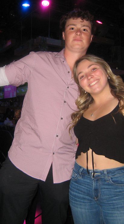 Two people posing and smiling in a neon-lit bar; man in a checkered shirt raising a red drink cup, woman in a black crop top and jeans leaning in on a nightclub dance floor.