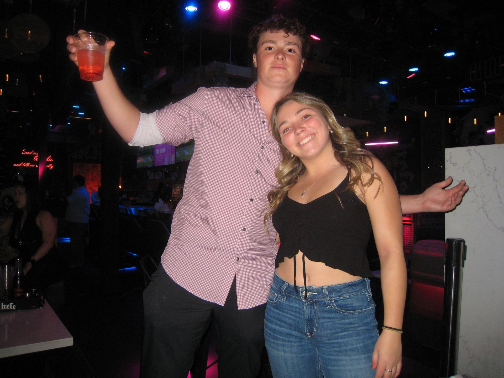 Two people posing and smiling in a neon-lit bar; man in a checkered shirt raising a red drink cup, woman in a black crop top and jeans leaning in on a nightclub dance floor.
