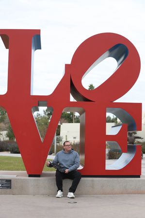 Person sitting on the base of a large red LOVE public art sculpture in an outdoor park, holding a black device beneath a cloudy sky