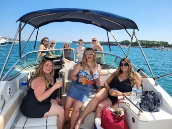Eight women in swimsuits and sunglasses smiling and holding drinks aboard a sunlit motorboat under a navy bimini, turquoise water and other boats visible on a clear summer day.